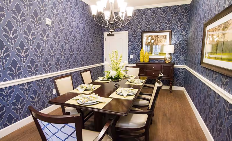 Formal dining room with a dark wood table set for six, patterned blue wallpaper, chandelier, and a sideboard with decor.