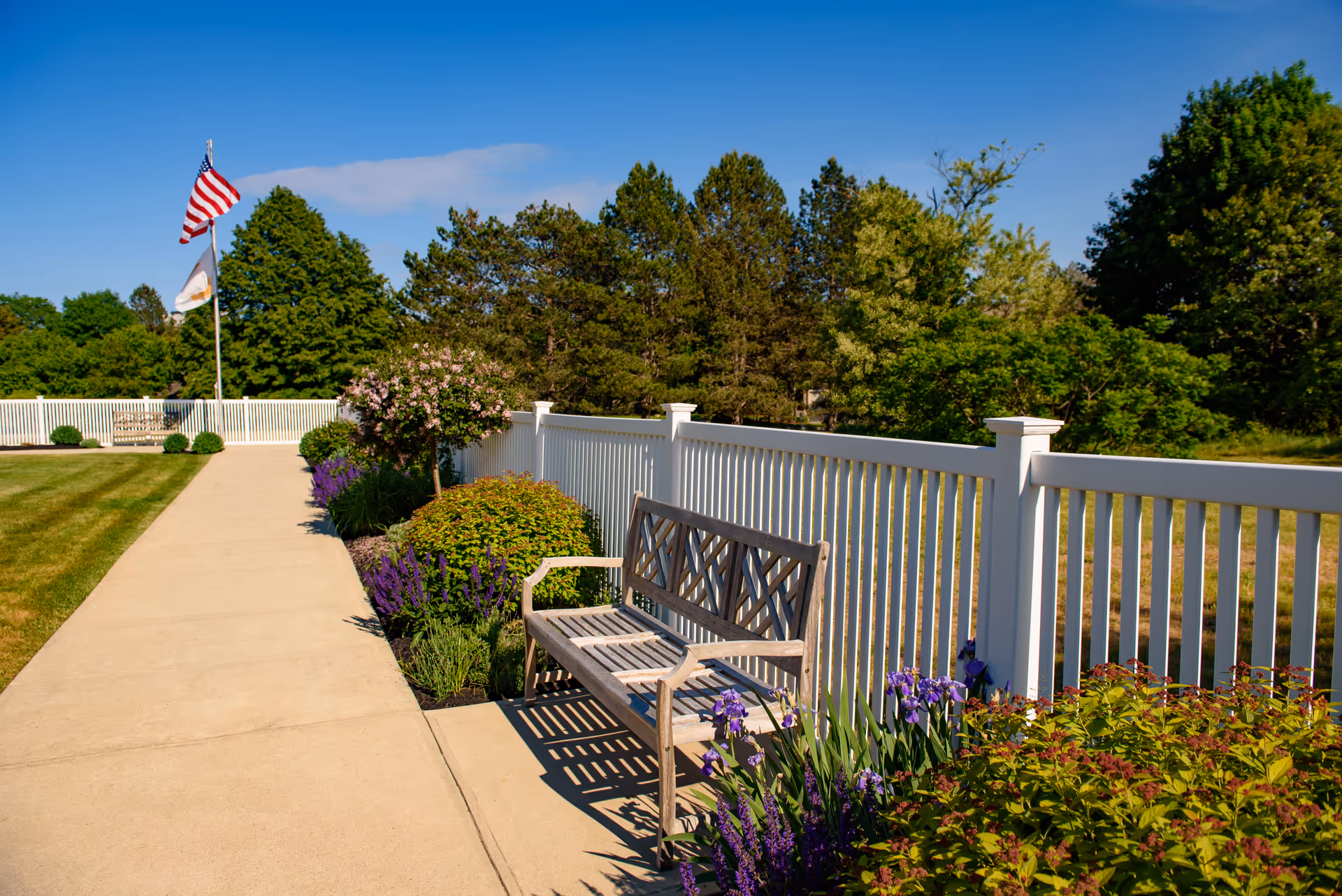 A wooden bench beside a paved walkway and white fence surrounded by flowering plants, trees, and flags under a blue sky.