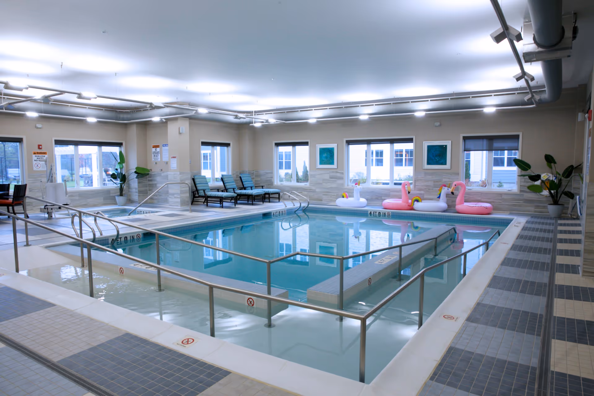 Indoor swimming pool area with a shallow pool section separated by a railing from a deeper pool. The pool has inflatable floaties shaped like unicorns and flamingos. There are lounge chairs along the far wall and large windows letting in natural light. The room has tiled floors and walls with modern lighting on the ceiling.