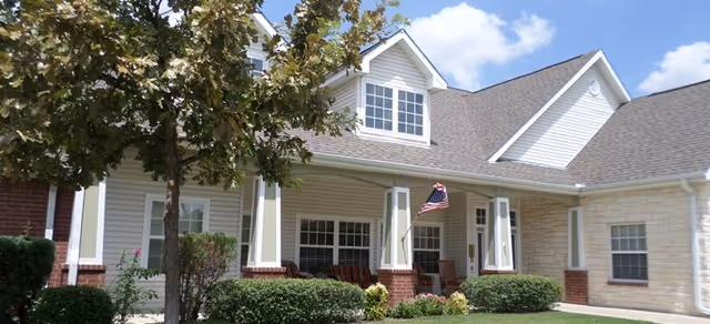 Front exterior of a residential building featuring a covered porch with rocking chairs, an American flag, and manicured shrubs.