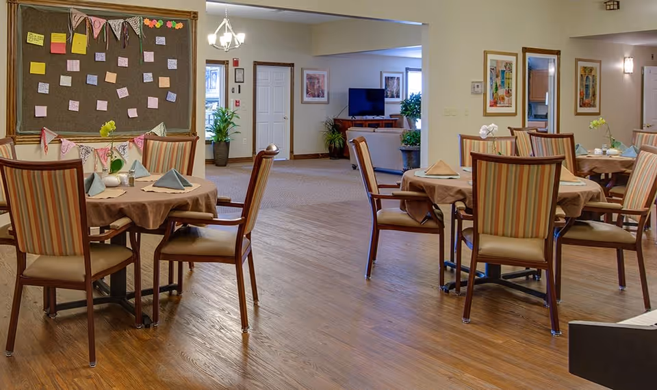 A dining area in a senior living facility with round tables covered with brown tablecloths and set with folded napkins. The chairs have striped upholstery. In the background, there is a bulletin board with colorful notes and decorations, and a living room area with a TV and plants is visible through an open doorway.