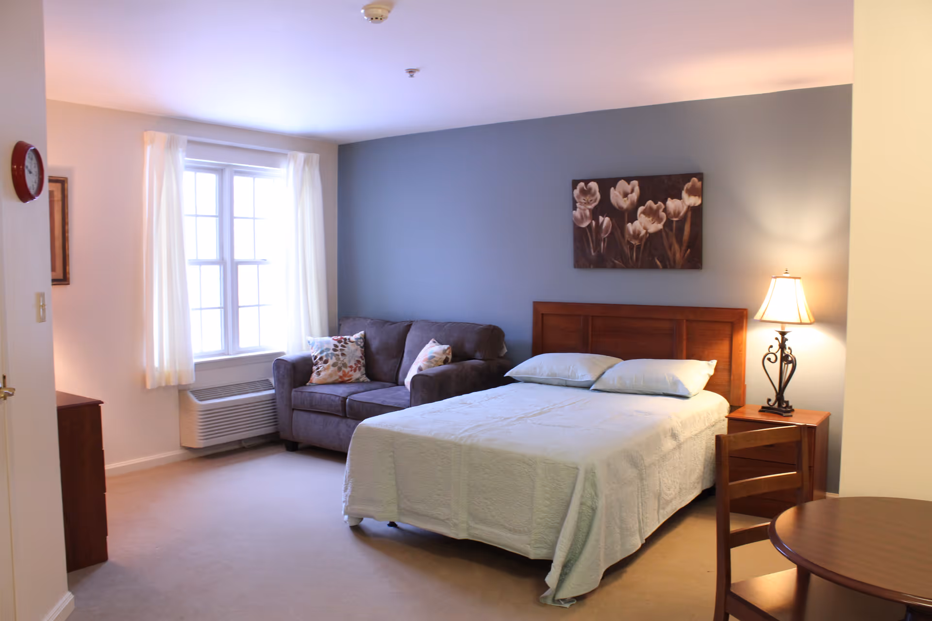 A well-lit bedroom with a double bed covered in a light-colored quilt, two pillows, a wooden headboard, and a bedside table with a decorative lamp. There is a gray couch with floral pillows near a window with white curtains. A round wooden table with a chair is partially visible in the foreground. The walls are painted light gray and white, and a floral painting hangs above the bed.
