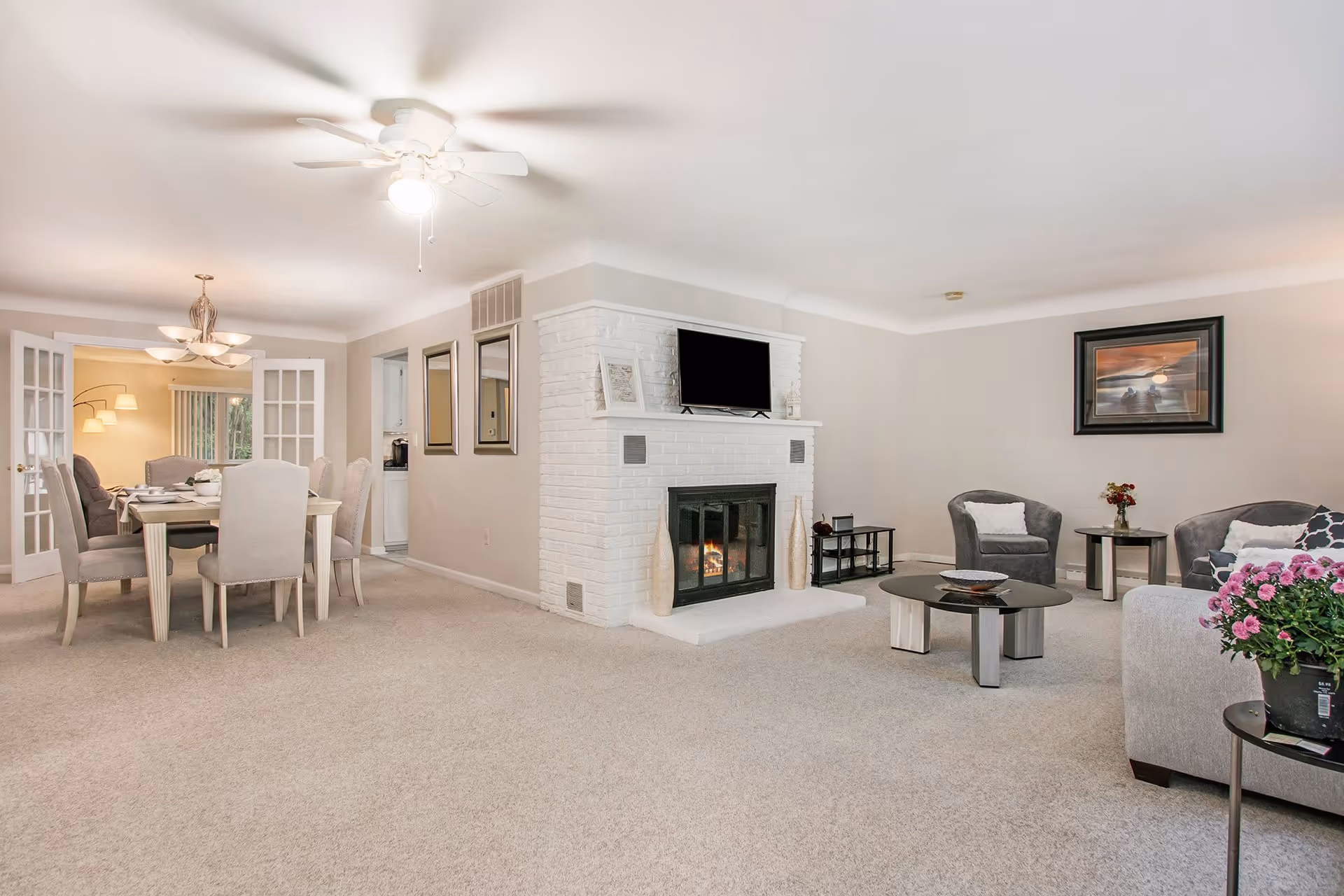 A spacious living room and dining area with beige carpet and light-colored walls. The living room features a white brick fireplace with a TV mounted above it, two gray armchairs, a gray sofa, a round coffee table, and a side table with a flower pot. The dining area has a rectangular dining table with six upholstered chairs and a chandelier overhead. There are two mirrors on the wall near the fireplace and a framed picture hanging on the living room wall.