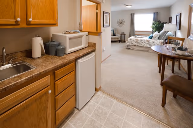 Interior view of a senior living facility unit showing a small kitchenette with wooden cabinets, a sink, a microwave, and a mini refrigerator. The kitchenette opens into a carpeted living and sleeping area with a bed, a chair, a small round dining table set for two, and a window with curtains letting in natural light.