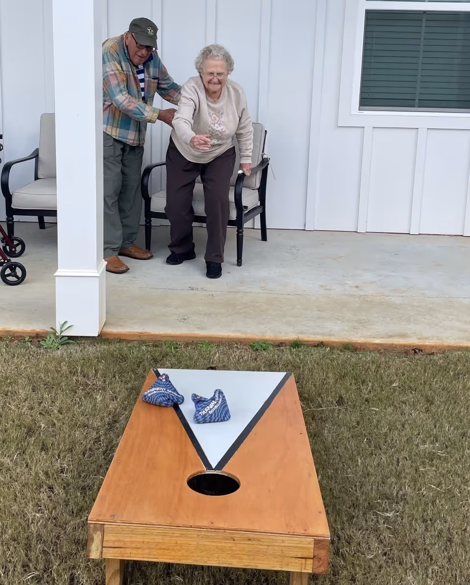 Two older adults stand on a covered porch tossing bean bags toward a wooden cornhole board on the lawn.