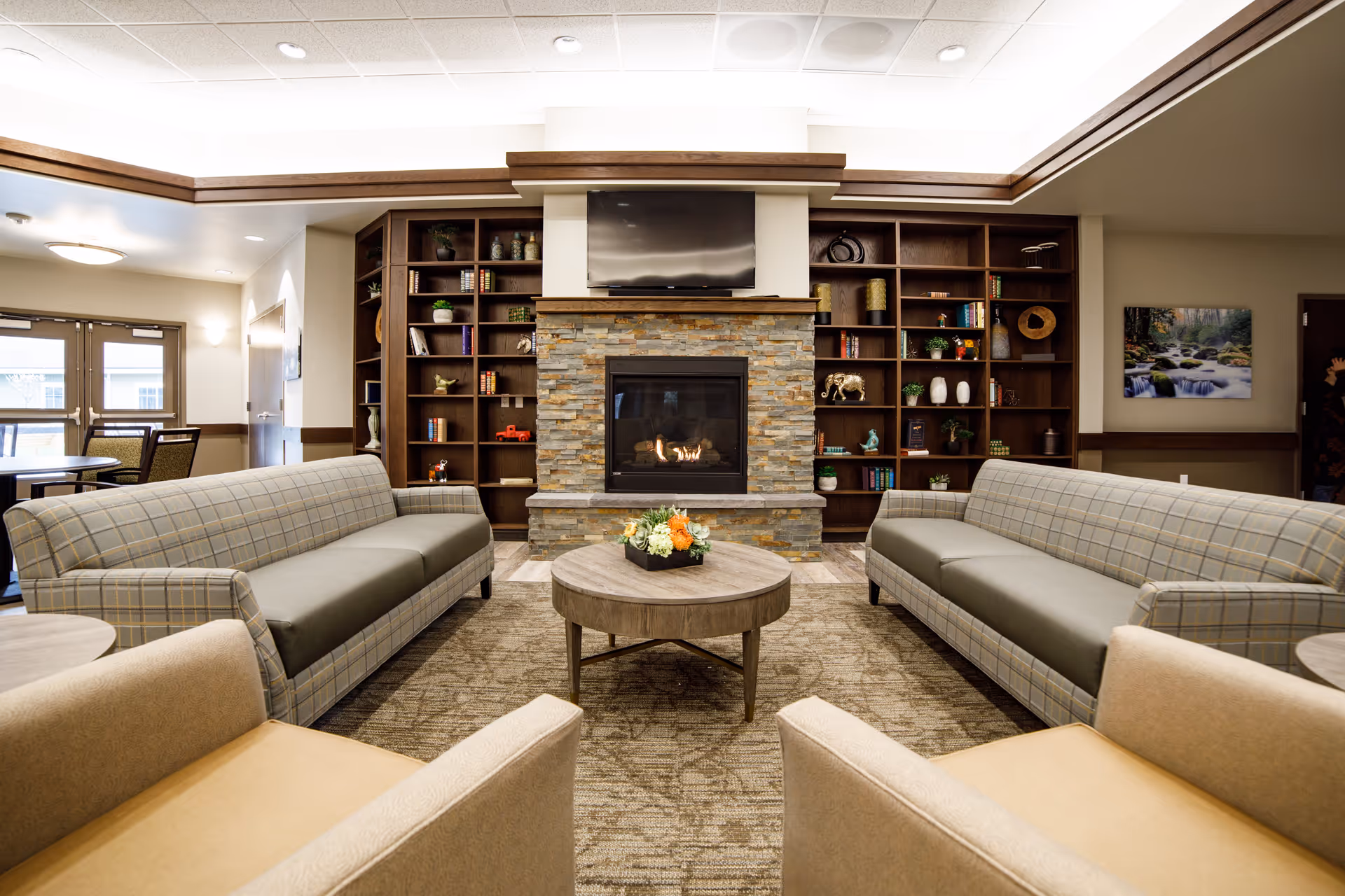 A cozy living room with two plaid sofas facing each other and two beige armchairs in the foreground. A round wooden coffee table with a floral arrangement is centered on a patterned carpet. The room features a stone fireplace with a TV mounted above it, flanked by built-in wooden bookshelves filled with books and decorative items. In the background, there is a dining area with tables and chairs near windows.
