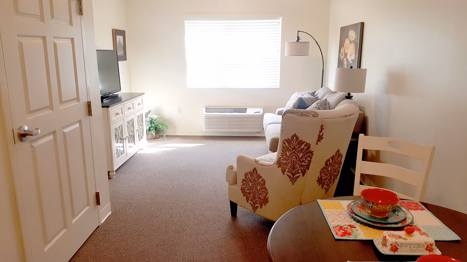 A bright living room with a window covered by blinds letting in natural light. The room features a patterned armchair, a sofa with cushions, a floor lamp, a TV on a white cabinet, a small plant in the corner, and a round dining table with a chair, placemats, and colorful dishes.