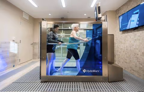 An elderly woman walking on an underwater treadmill inside a HydroWorx aquatic therapy tank, assisted by a younger woman in a modern indoor therapy or rehabilitation room.