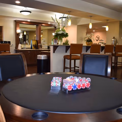 A room with a round poker table in the foreground holding poker chips and playing cards. In the background, there is a bar area with high chairs, a countertop with flower arrangements, and pendant lights hanging from the ceiling.