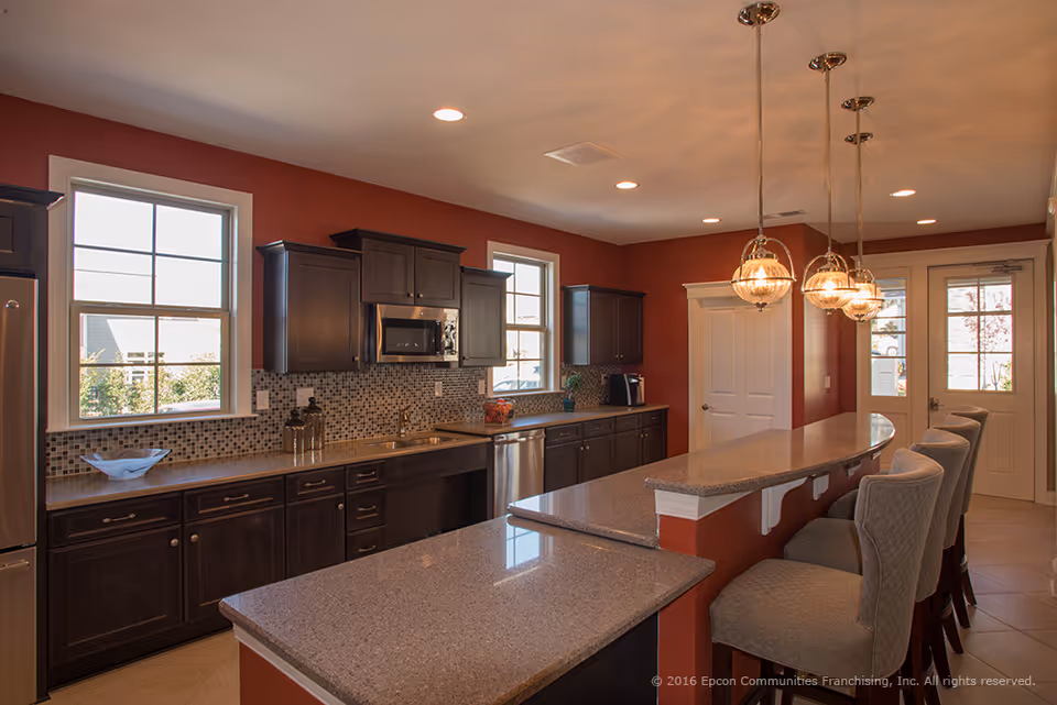 A modern kitchen with dark wood cabinets, granite countertops, and a mosaic tile backsplash. The kitchen features stainless steel appliances including a refrigerator, microwave, and dishwasher. There are three pendant lights hanging above a breakfast bar with four upholstered chairs. Two windows provide natural light, and a door with glass panels is visible in the background.