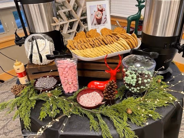 A festive hot chocolate station with two large beverage dispensers, a platter of assorted cookies, jars filled with pink and green candy, bowls of chocolate chips and crushed peppermint, decorated with pinecones, greenery, and a red reindeer figurine on a black tablecloth.
