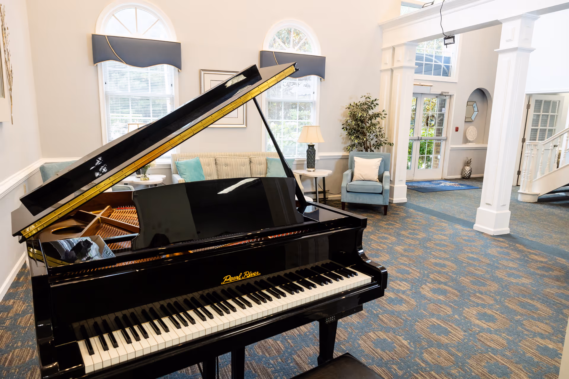 Interior view of a senior living facility lounge area featuring a black Pearl River grand piano in the foreground. Behind the piano, there is a beige sofa with blue cushions, a blue armchair with a white pillow, a small round side table with a lamp, and large arched windows with blue valances. The room has patterned blue carpet, white walls with molding, and a doorway leading to another area with stairs and glass doors.