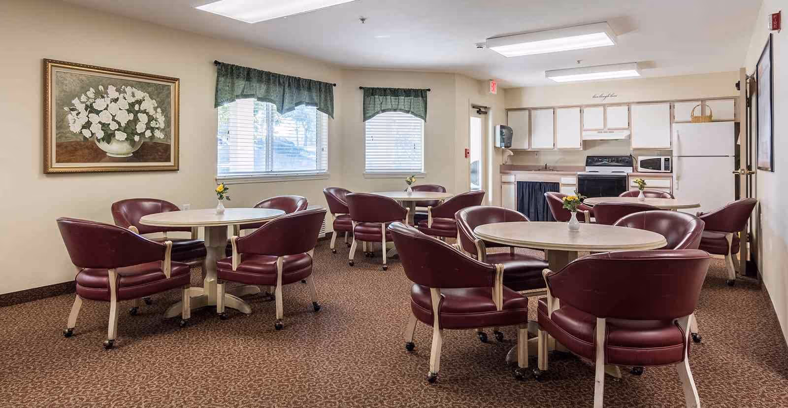 A communal dining area with several round tables, each surrounded by maroon cushioned chairs on wheels. The room has beige walls, two windows with green valances, a painting of white flowers on the wall, and a kitchen area in the background with white cabinets, a stove, microwave, and refrigerator.