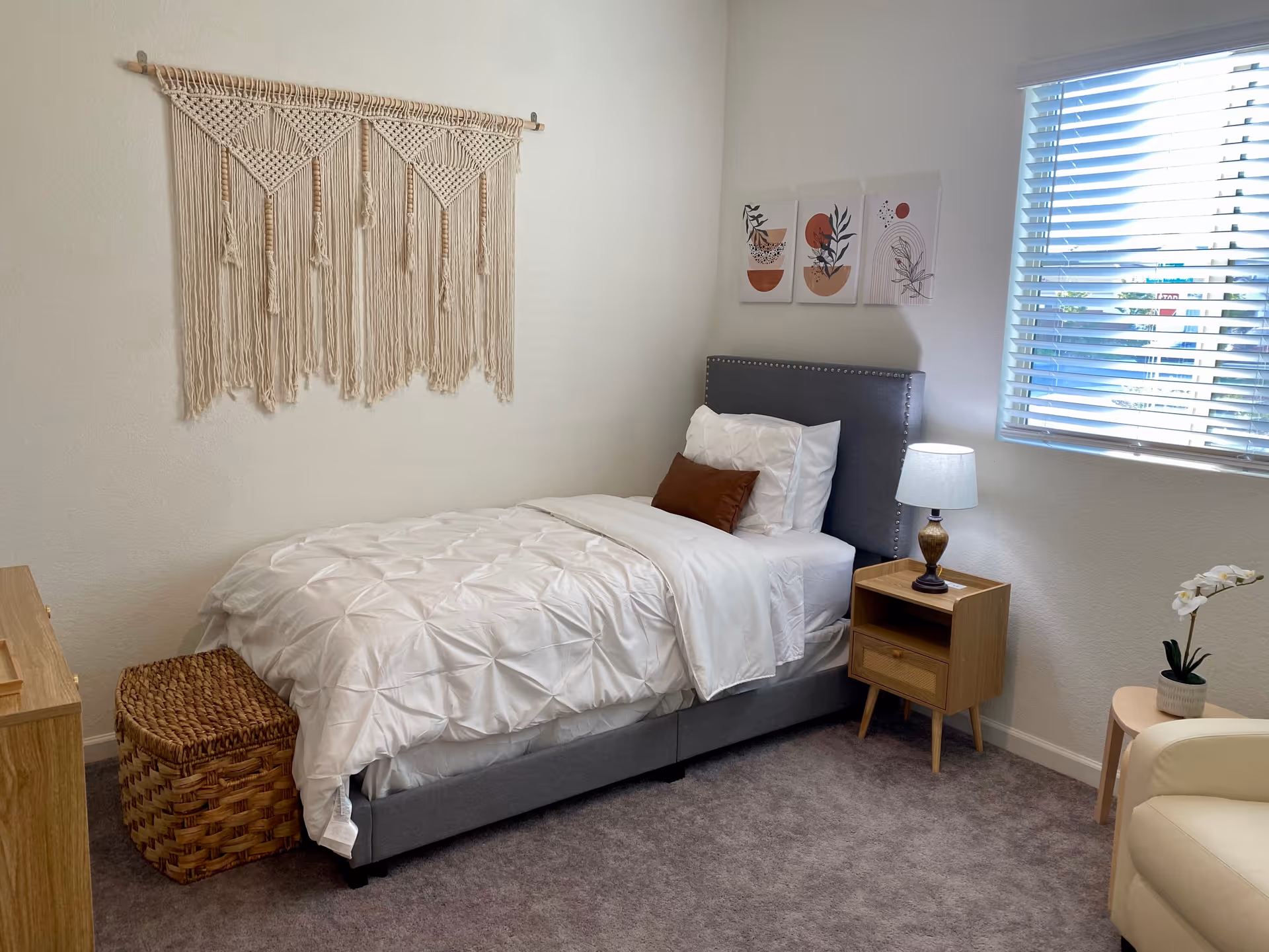 A cozy bedroom with a single bed featuring white bedding and a brown accent pillow. There is a wooden nightstand with a lamp beside the bed, a wicker basket at the foot of the bed, and a beige armchair partially visible. The walls are decorated with a macrame wall hanging and three abstract art prints. A window with blinds allows natural light into the room.