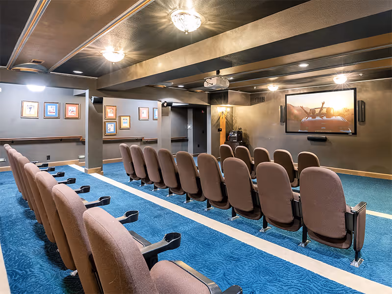 Carpeted community theater room with rows of upholstered seats facing a large wall-mounted screen and ceiling projector.