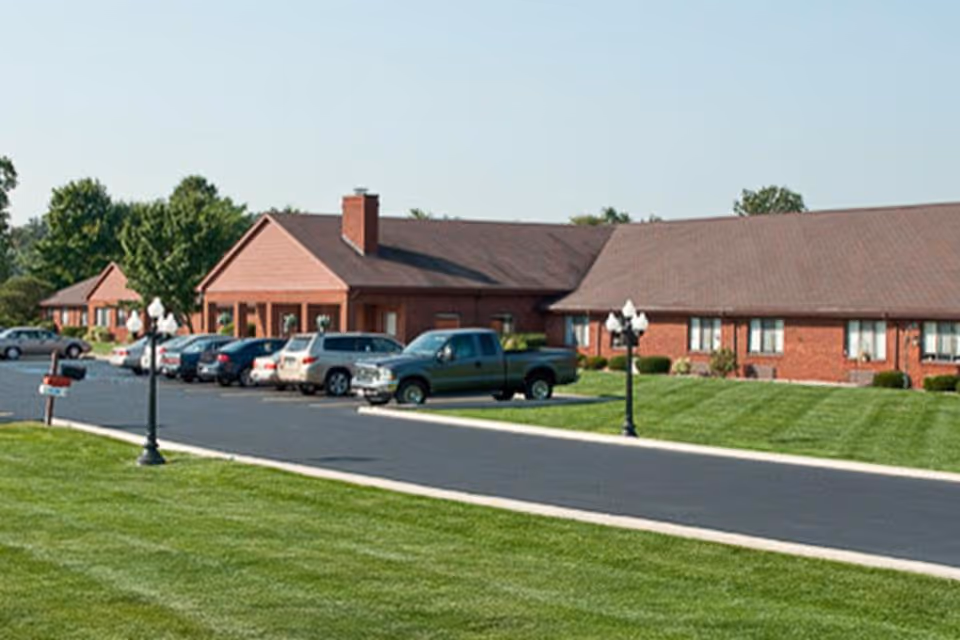 Single-story brick care facility building with a row of parked cars, a driveway and neatly mowed lawns in front.