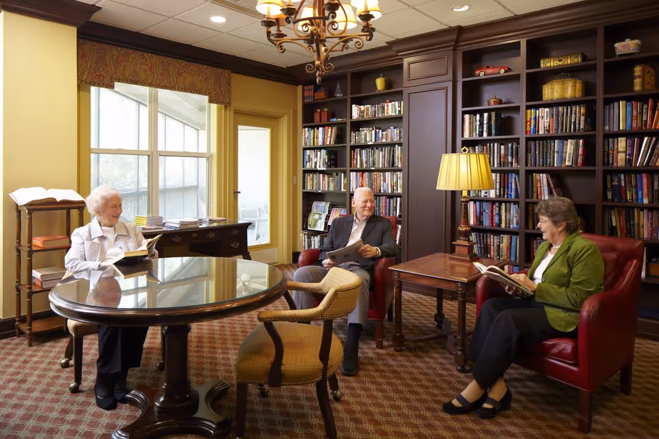 Three elderly people sitting and reading in a cozy library room with wooden bookshelves filled with books, a round glass-top table, a wooden side table with a lamp, and large windows letting in natural light.