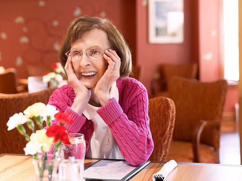An elderly woman wearing glasses and a pink sweater sits at a table in a warmly decorated dining area, smiling and resting her face in her hands. On the table are a glass of water, a menu, a napkin with cutlery, and a small vase with red and white flowers. The background shows upholstered chairs and a softly lit room with red walls.