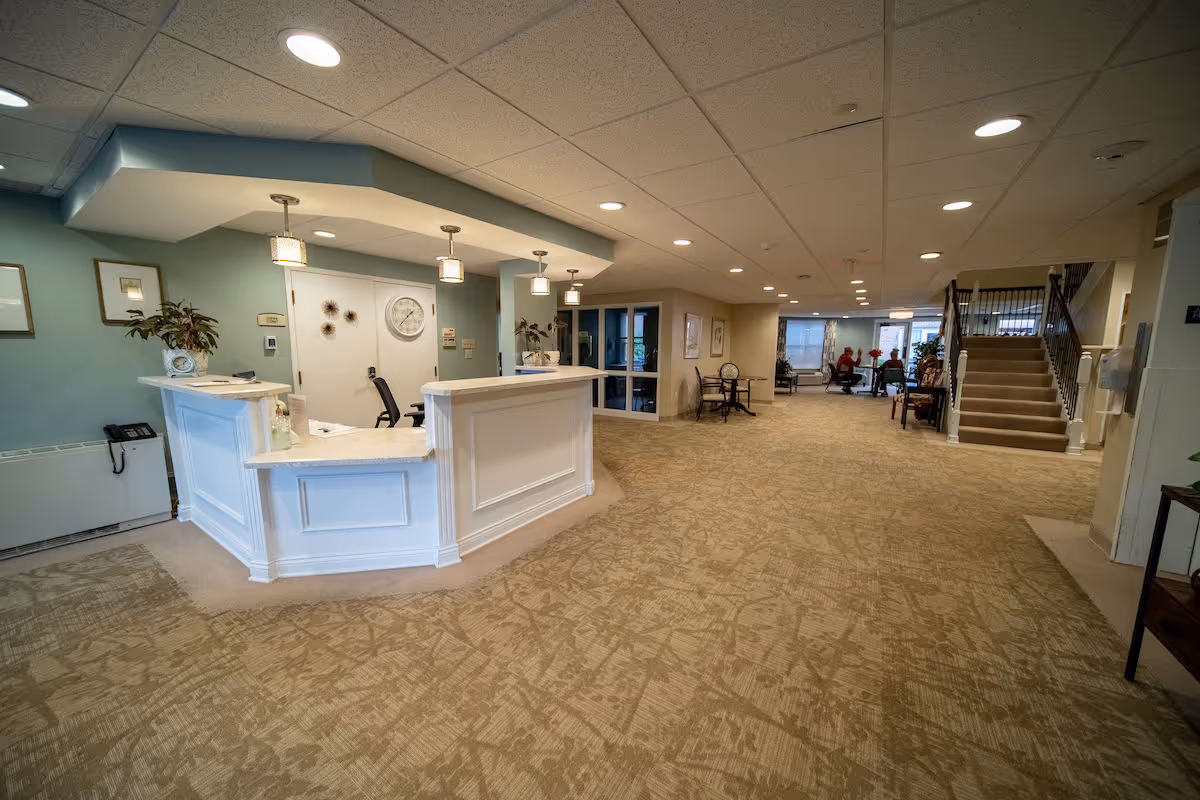 Reception desk and spacious lobby of a senior living facility with seating and a staircase in the background.
