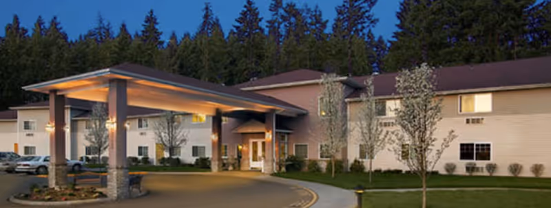 Exterior view of a senior living facility building at dusk with a covered entrance, several windows, small trees, and a parking area with cars.