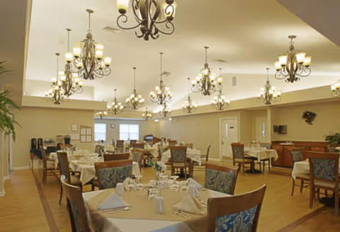 A spacious dining room with multiple tables covered with beige tablecloths, each set with folded napkins, glasses, and silverware. The room features wooden chairs with patterned cushions, several ornate chandeliers hanging from the ceiling, and large windows letting in natural light. The walls are light-colored, and there are plants and framed items on the walls.