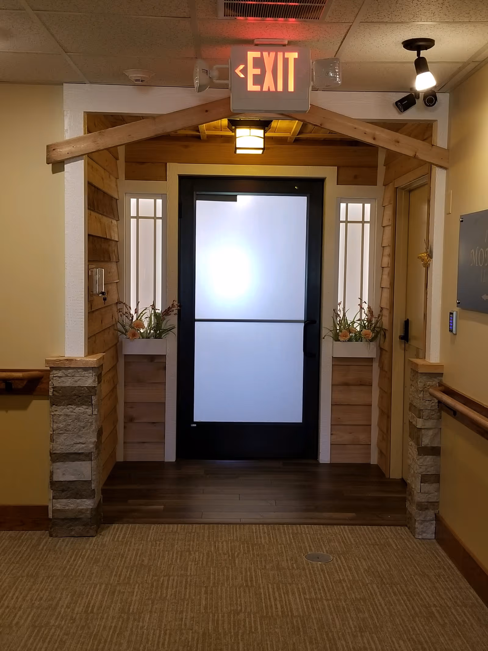 Interior hallway of a retirement community with a frosted glass door at the end, flanked by wooden paneling and flower arrangements. Above the door is an illuminated exit sign with emergency lights. The hallway has carpeted flooring and handrails on both sides.