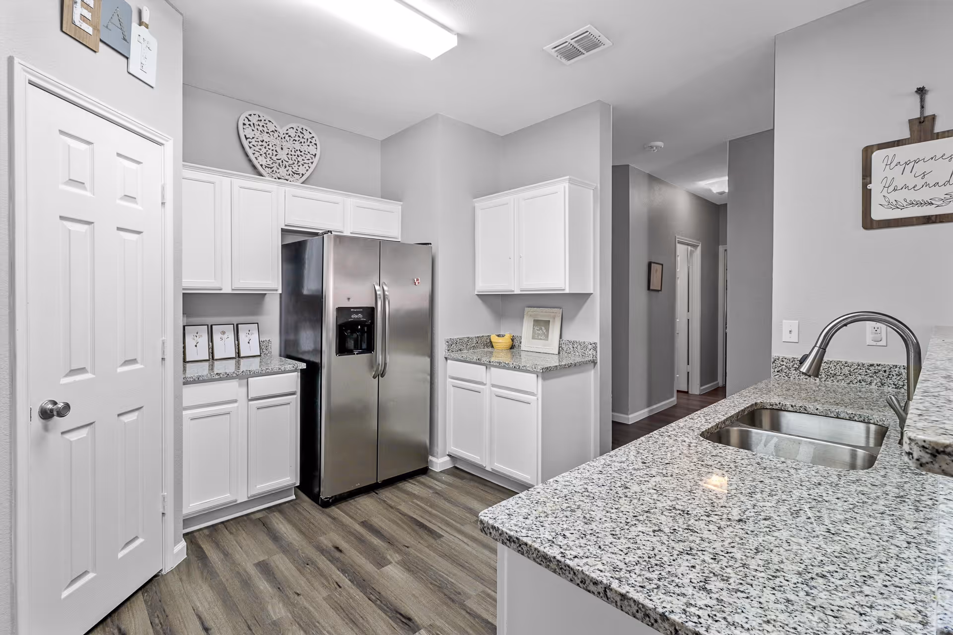 Modern kitchen with white cabinets, granite countertops, a stainless steel refrigerator, and a double sink. The floor is wood-style laminate, and there are decorative items such as framed pictures and a heart-shaped wall decoration.