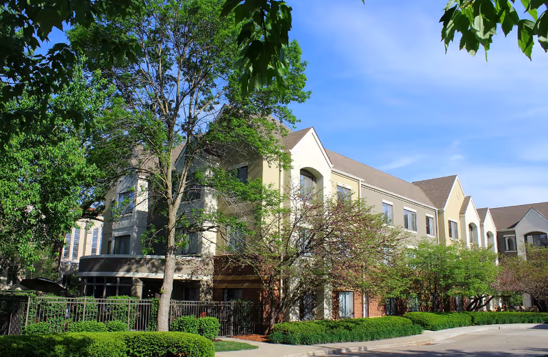 Exterior view of a multi-story senior living facility building surrounded by green trees and shrubs under a clear blue sky.