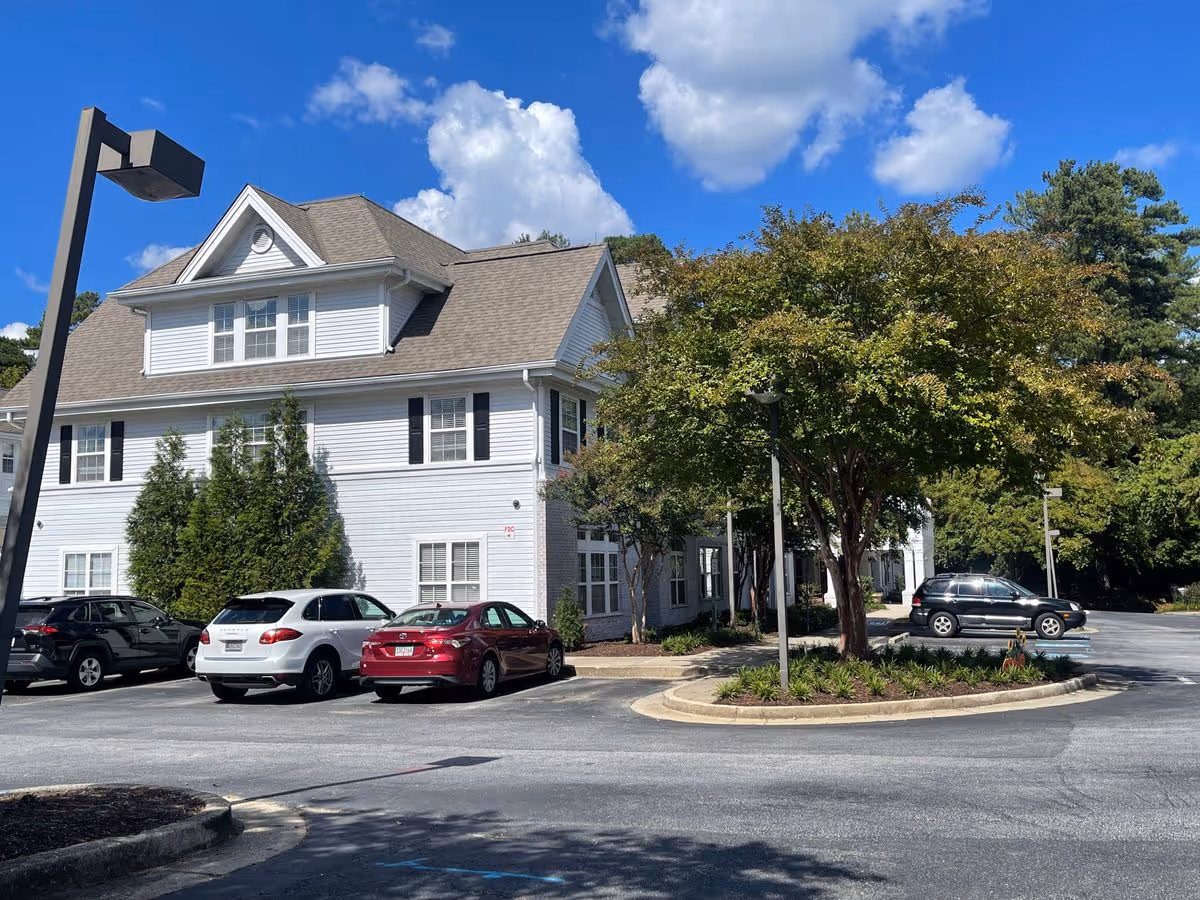 White multi-story senior living building with parked cars and a landscaped parking lot under a blue sky.