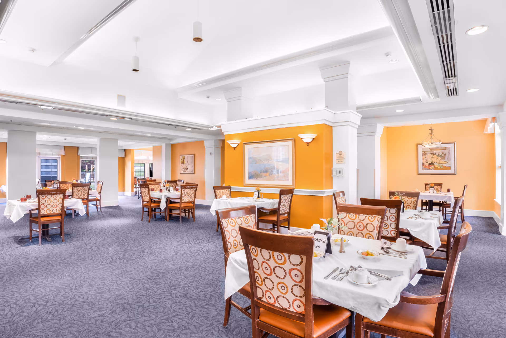 Bright dining room with orange-accented walls and multiple tables set with white tablecloths and patterned chairs.