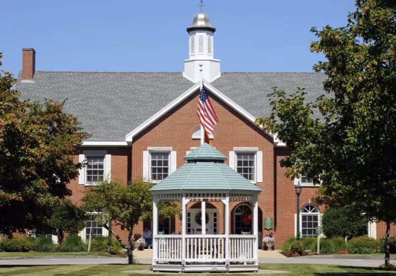 Front view of a brick senior living building with a white gazebo and an American flag in the foreground.