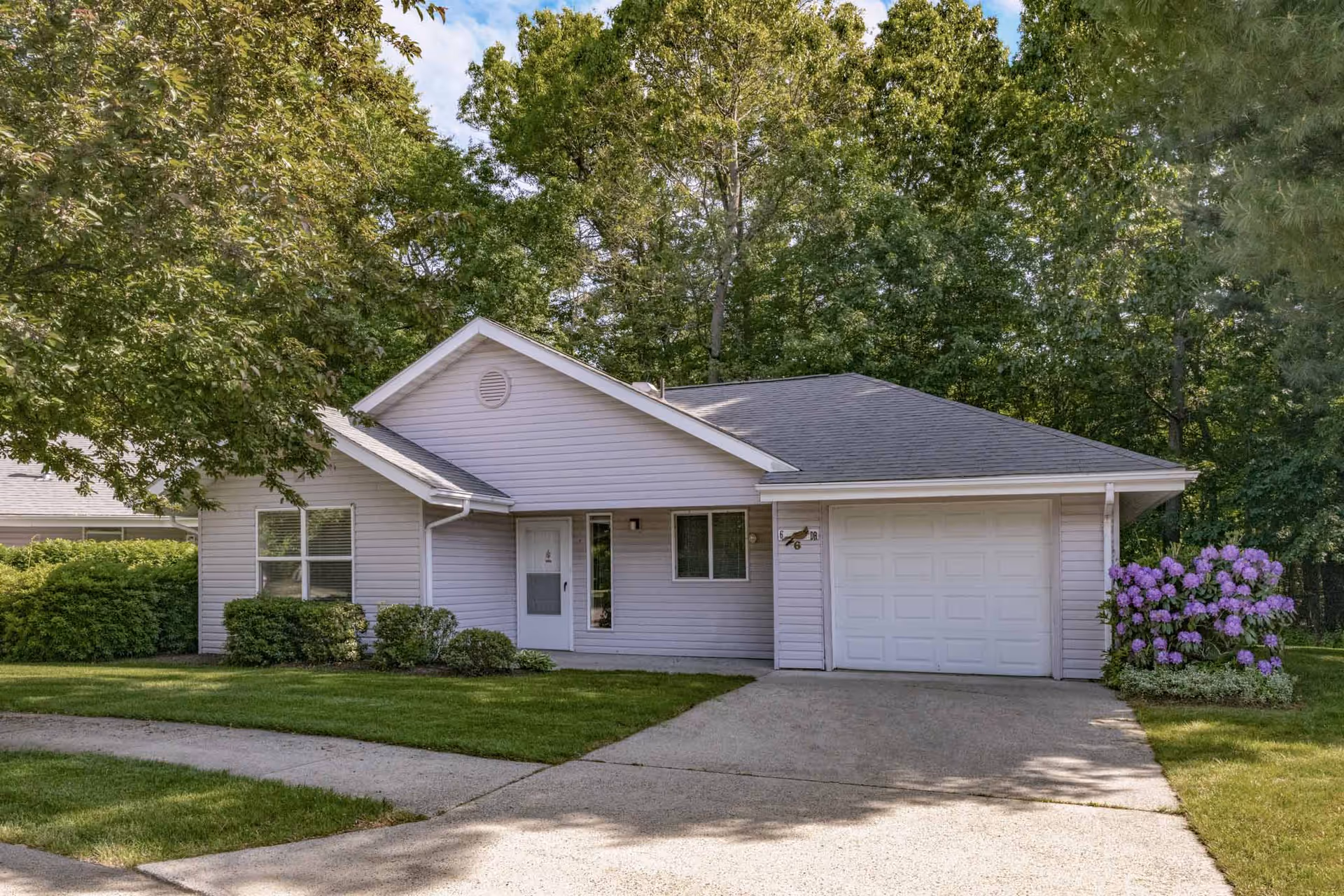 Single-story house with light gray siding and a dark gray roof, featuring a white front door and a single-car garage. The house is surrounded by green grass, bushes, and trees, with a concrete driveway leading up to the garage. Purple flowers are blooming near the garage on the right side.