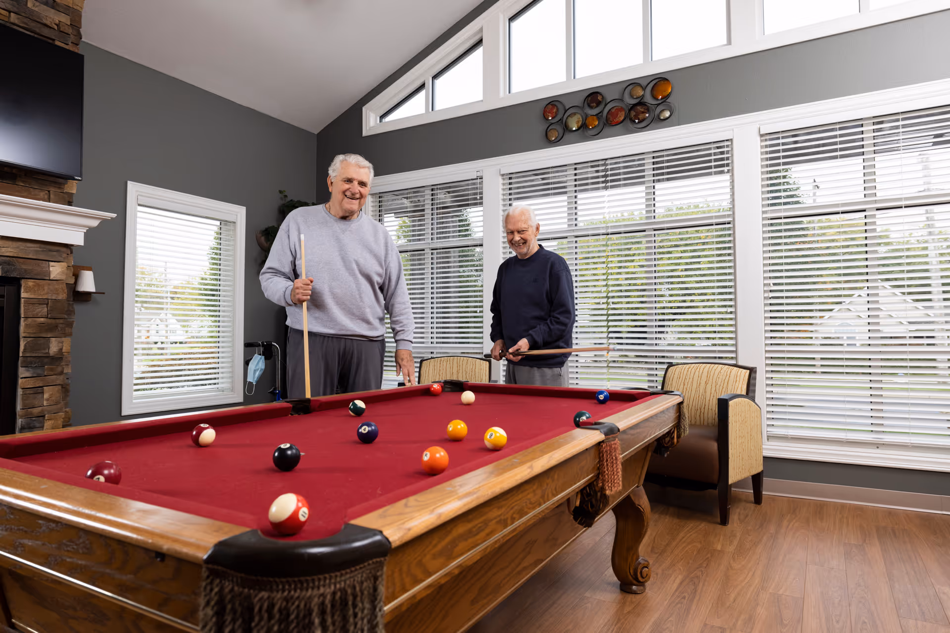Two elderly men playing pool on a red felt pool table in a bright room with large windows and wooden flooring. The room has a stone fireplace, a mounted TV, and comfortable chairs near the windows.