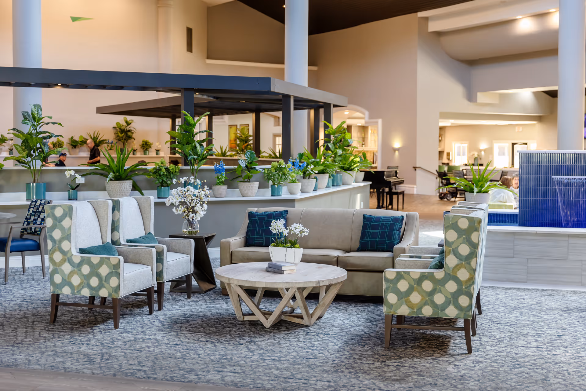 A spacious and well-lit senior living facility common area with comfortable seating including a beige sofa, patterned armchairs, and a round wooden coffee table with a small plant and books. The area is decorated with numerous potted plants on a raised ledge behind the seating. In the background, there are more seating areas, a piano, and large white columns supporting the high ceiling.