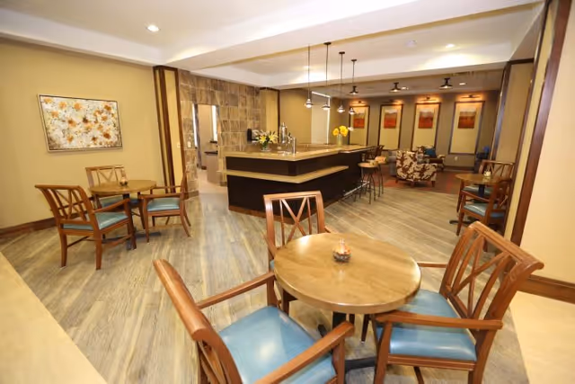 Interior view of a senior living facility common area featuring wooden round tables with blue cushioned chairs, a central bar or counter with pendant lighting, and a seating area with armchairs and framed artwork on the walls.