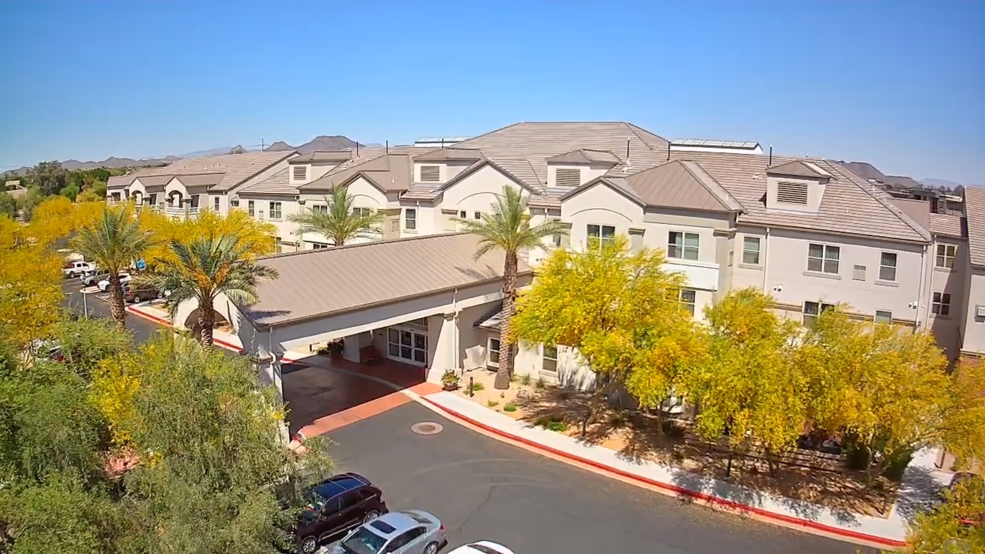 Exterior view of Arrowhead Valley Retirement Resort showing a large, multi-story building with a covered entrance, surrounded by palm trees and other greenery under a clear blue sky. Several cars are parked near the entrance and mountains are visible in the background.