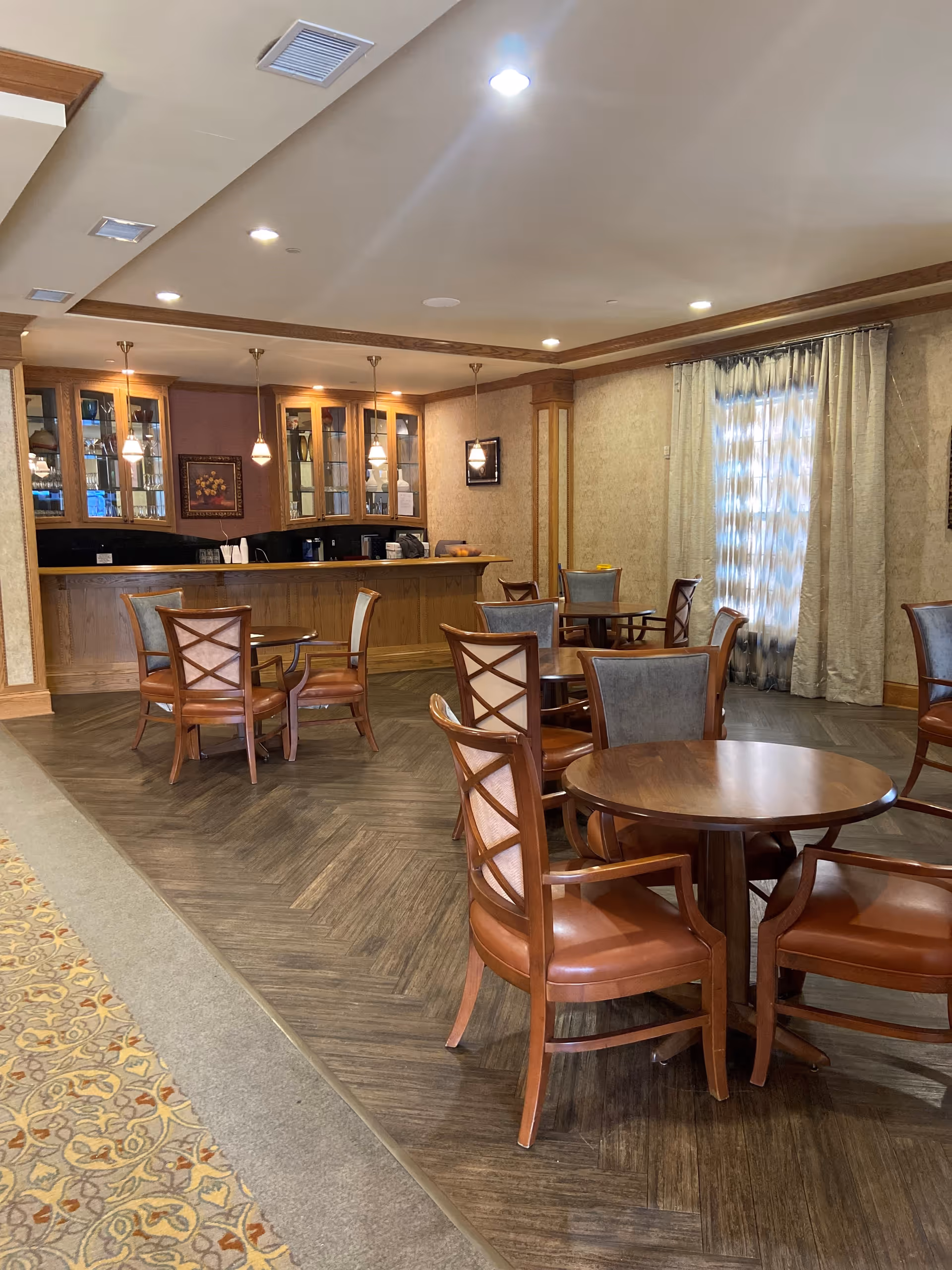 Interior view of a common area with multiple round wooden tables and chairs with leather seats. In the background, there is a wooden bar with glass cabinets and hanging pendant lights. The room has a patterned carpet and wood flooring, beige walls, and a window with curtains.