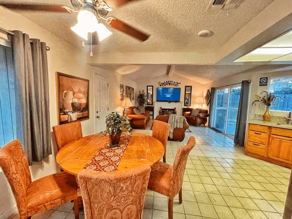Interior view of a senior living facility showing a dining area with a round wooden table and six upholstered chairs. A ceiling fan with lights is above the table. Beyond the dining area is a living room with leather armchairs, a fireplace, and a wall-mounted TV. Large sliding glass doors provide natural light and a view of the outdoors. The kitchen area with wooden cabinets and a countertop is visible to the right.
