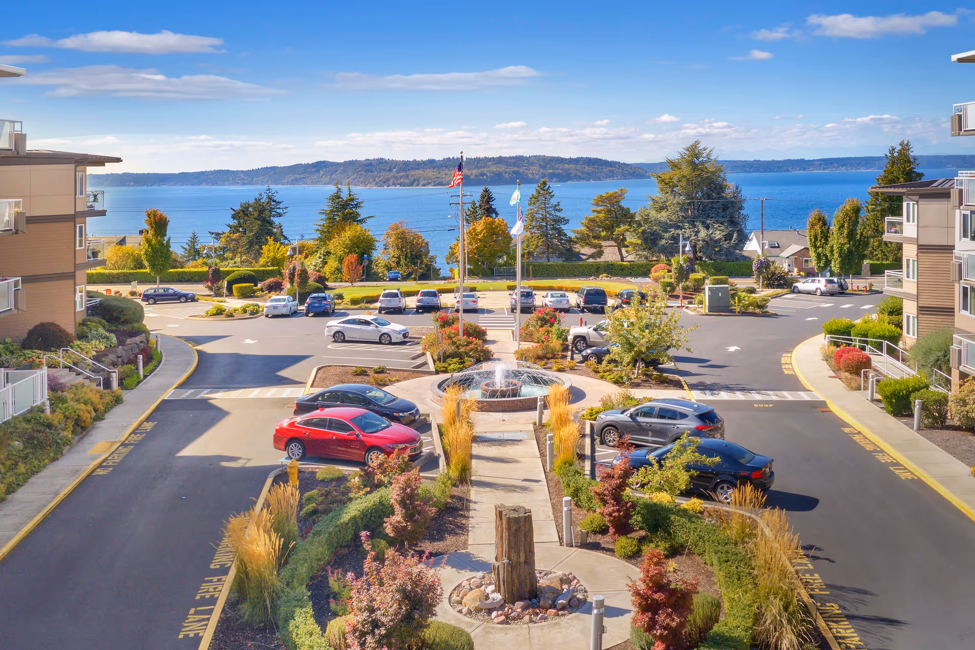 View of a parking lot with several cars parked around a central fountain and landscaped garden area, flanked by two multi-story buildings. In the background, there is a scenic view of a large body of water with hills on the horizon under a partly cloudy blue sky.