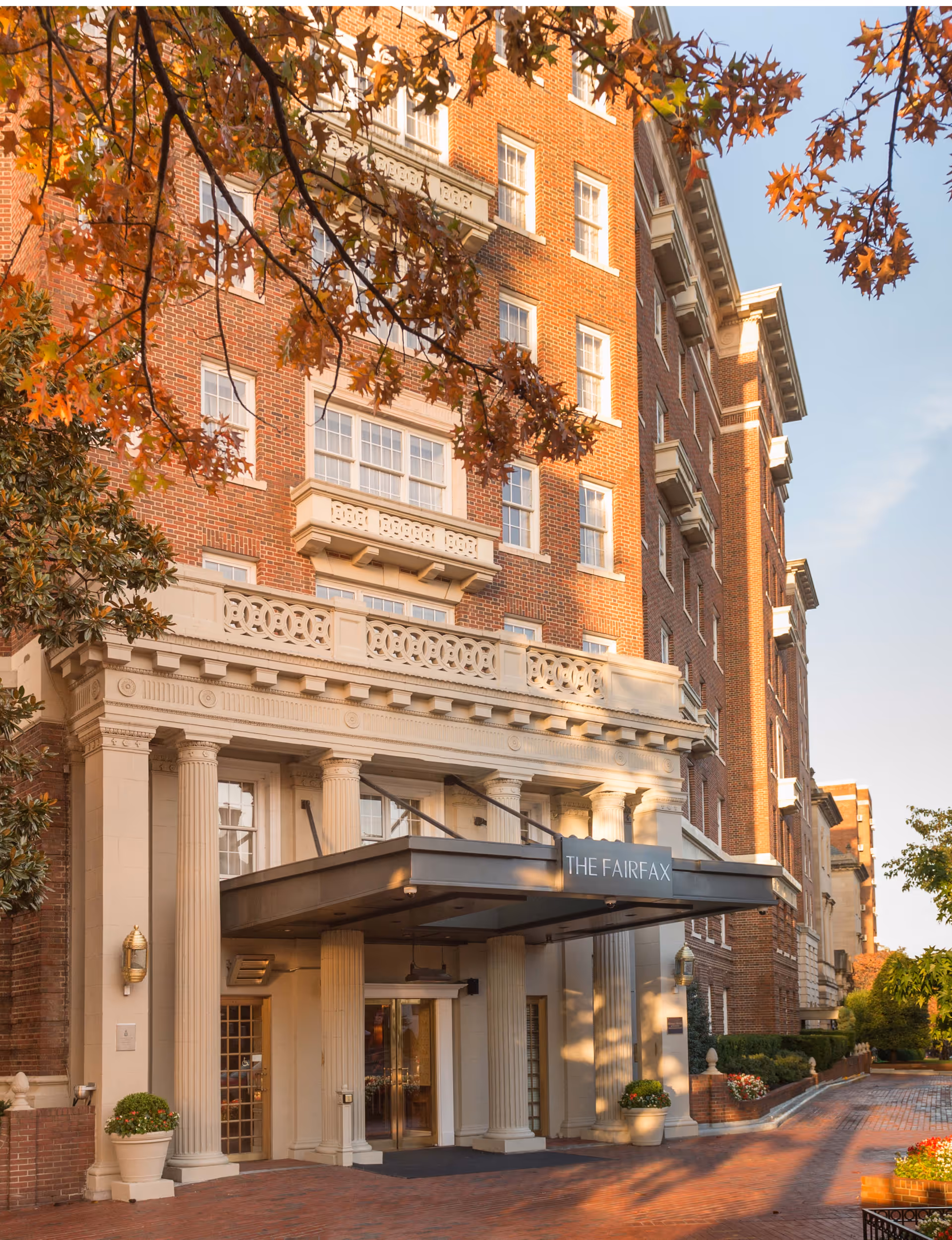 Exterior view of a multi-story brick building with classical architectural elements including columns and decorative railings. The entrance features a canopy with the sign 'THE FAIRFAX'. Trees with autumn-colored leaves frame the building.
