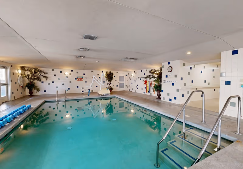 Indoor swimming pool with tiled walls featuring blue and gray square accents. The pool area includes metal handrails, steps leading into the water, a pool lift for accessibility, and pool noodles stacked on the side. There are potted plants and wall-mounted lights around the pool area.