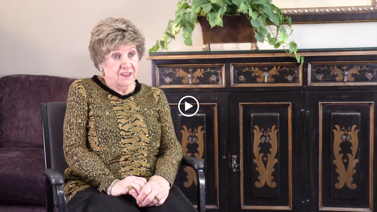 An elderly woman with short curly hair wearing a patterned sweater is seated in a chair in a living room. Behind her is a dark wooden cabinet with decorative floral designs and a green leafy plant on top. A purple couch is partially visible to the left.