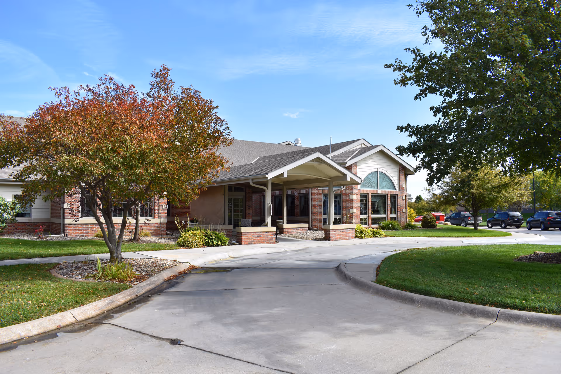Exterior view of Hillcrest Silver Ridge facility showing a brick building with a covered entrance, surrounded by green lawns and trees with autumn foliage under a clear blue sky.