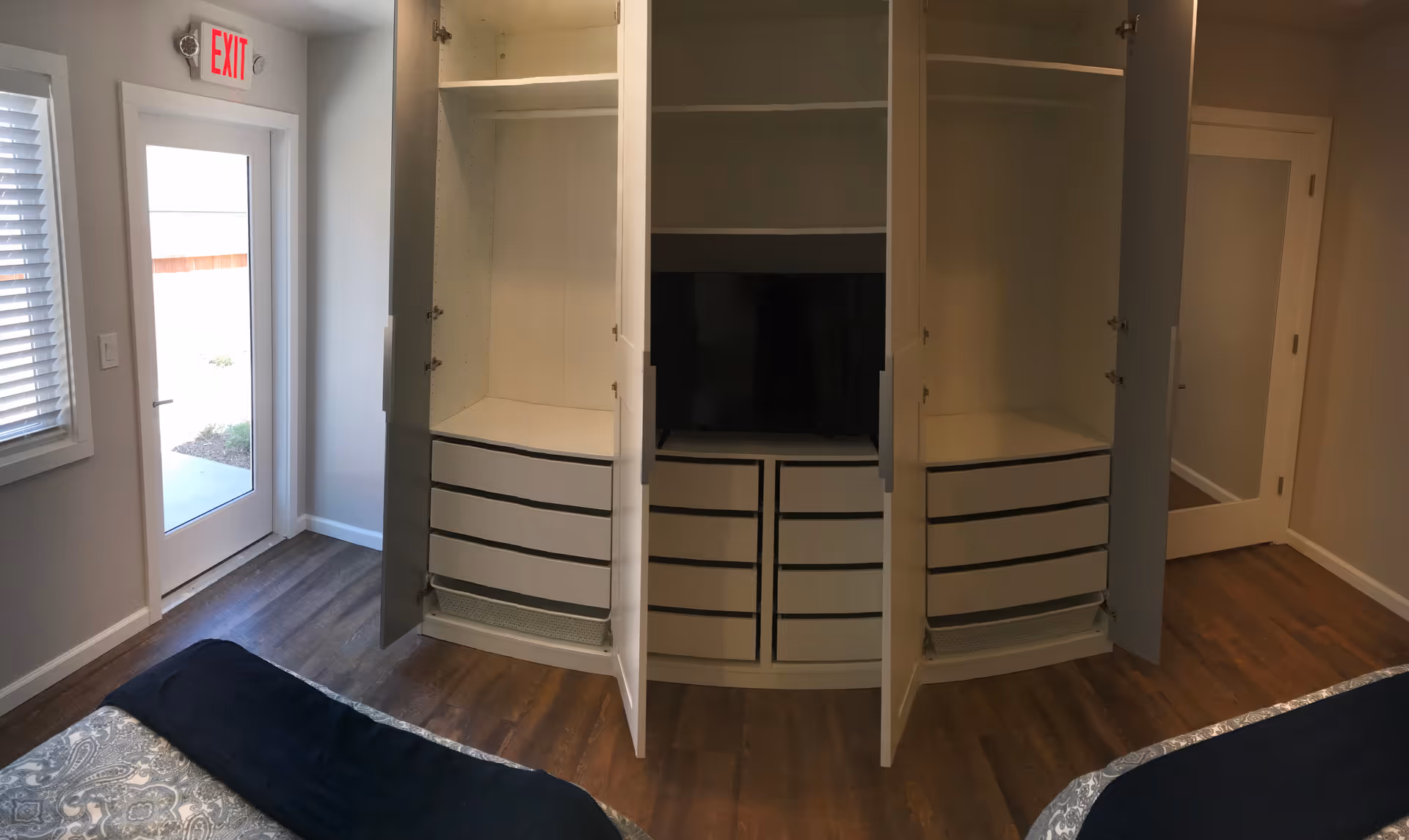 Bedroom interior with open built-in wardrobes and drawers, a bed edge in the foreground, and a glass exit door to the left.