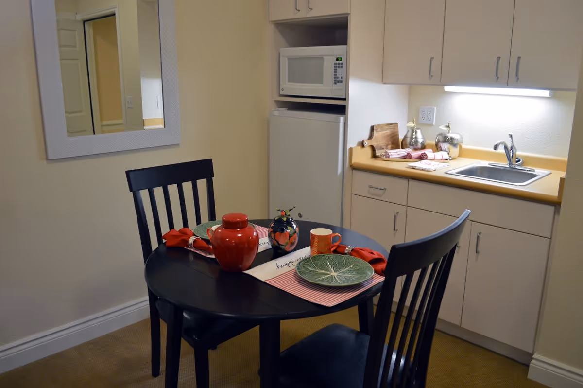 Small kitchen and dining nook with a round black table set for two in front of a kitchenette with sink, microwave, and cabinets.