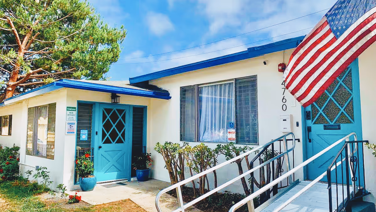 Exterior view of Hawthorne Terrace Care Home showing a single-story building with white walls and blue trim. There are two blue doors, one with a ramp and handrails leading up to it, and an American flag hanging near the entrance. Shrubs and potted plants are visible around the building under a partly cloudy sky.