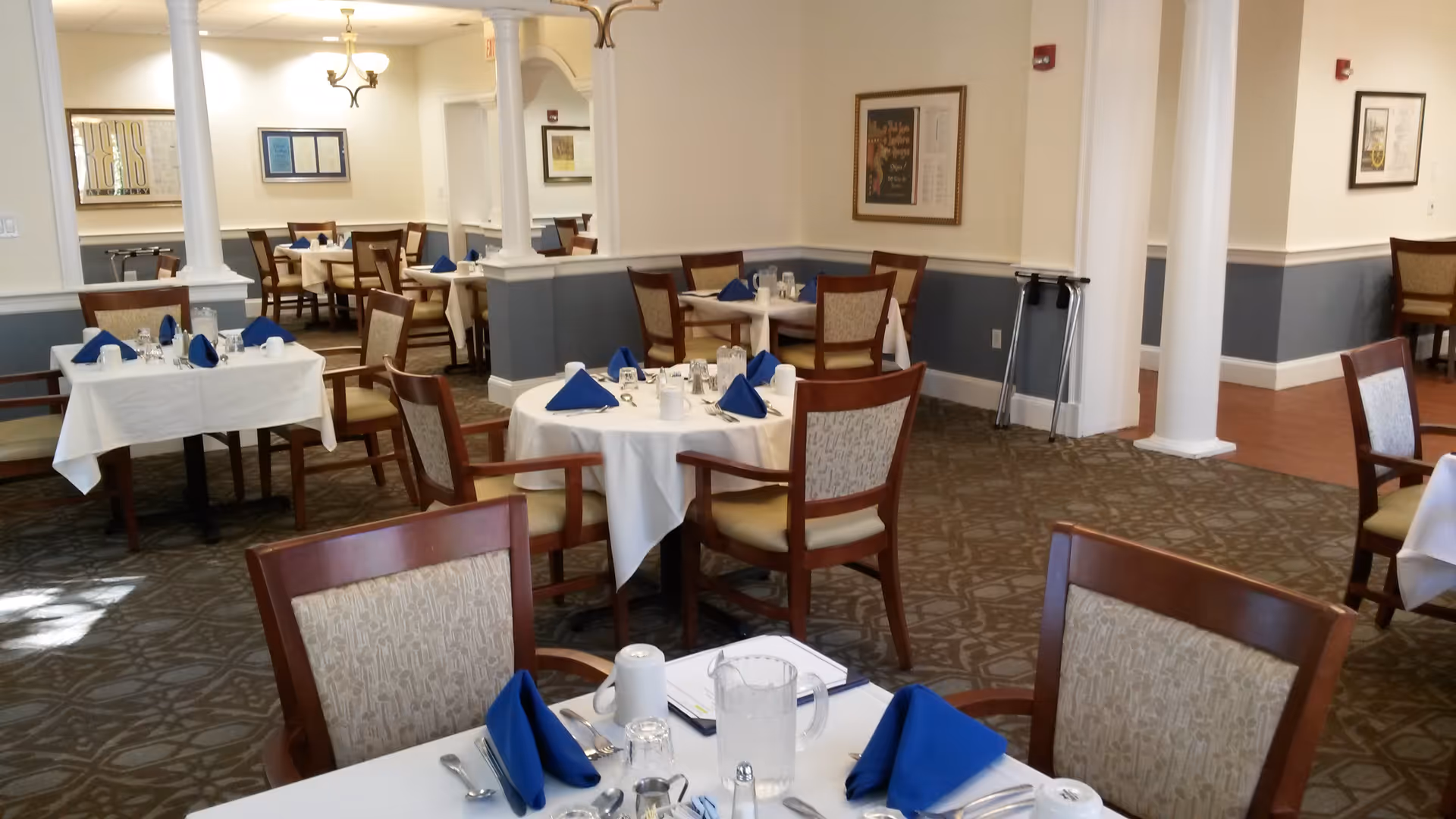Dining room with multiple round and rectangular tables set with white tablecloths and blue napkins.