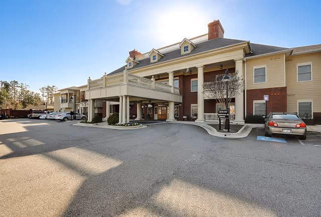 Exterior view of a two-story senior living facility building with a covered entrance, several parked cars, and a clear blue sky.