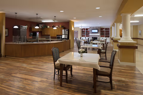 Interior view of a senior living facility dining area with wooden floors, tables covered with white tablecloths, chairs, and a kitchen area in the background. The space is well-lit with ceiling lights and has a warm, inviting atmosphere.