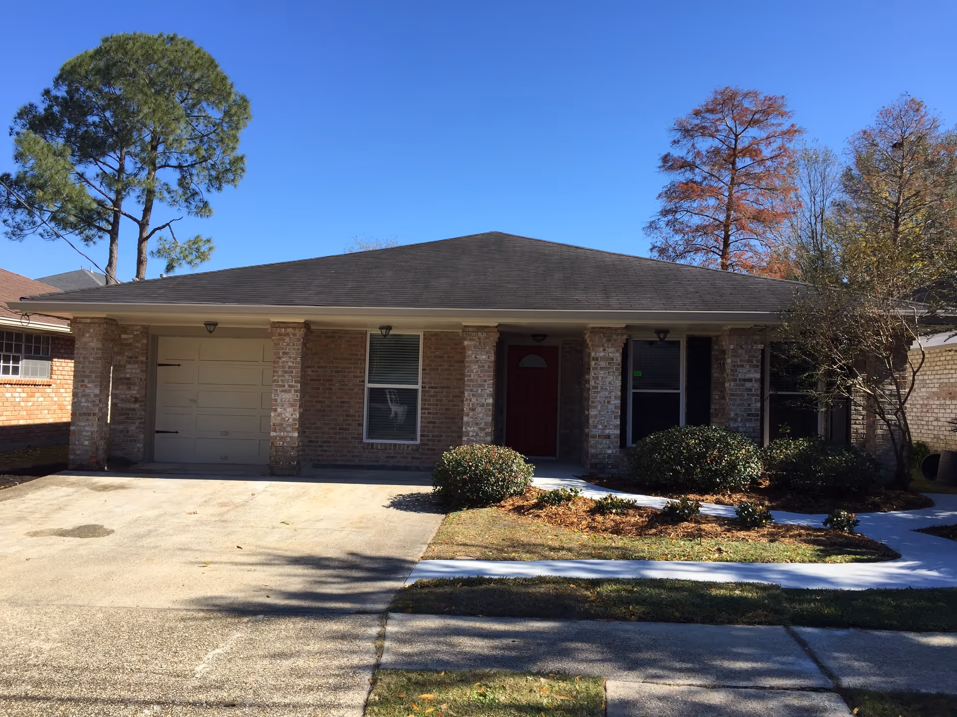 Single-story brick house with a dark shingled roof, a garage on the left, a red front door in the center, and several windows. The front yard has trimmed bushes and a concrete driveway and walkway. Tall trees are visible in the background under a clear blue sky.